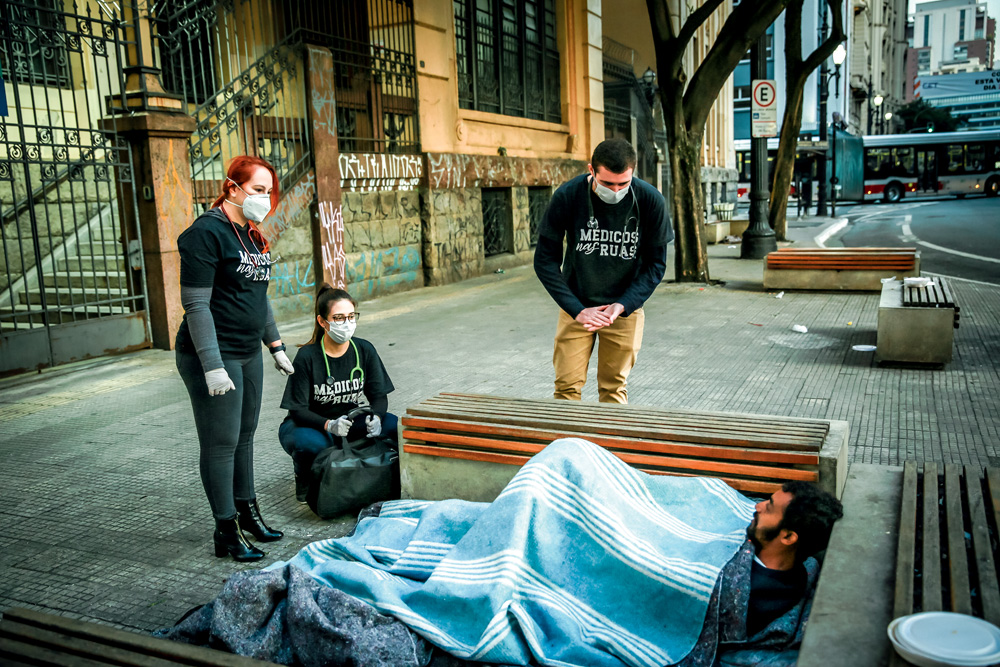 Um homem e duas mulheres, todos brancos, atendem um homem deitado no chão, em situação de rua. Eles usam um uniforme preto e possuem estetoscópios no pescoço