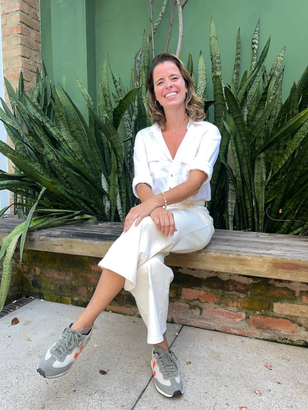 Renata Salles, do Open Air, posa de roupa branca em jardim, com as pernas cruzadas e sorrindo.