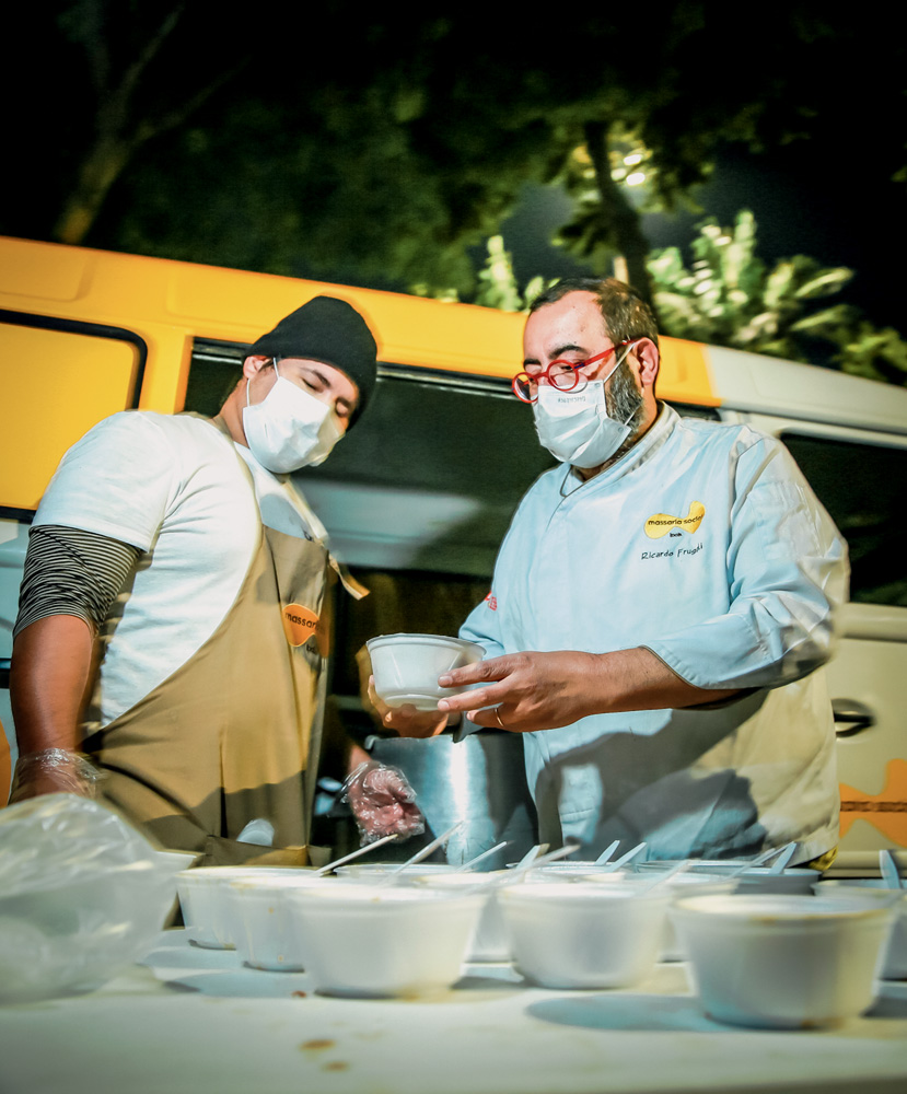 Dois homens, um deles com uma dólmã e o outro com um avental de cozinha, preparam sopas em uma mesa ao lado de uma van amarela