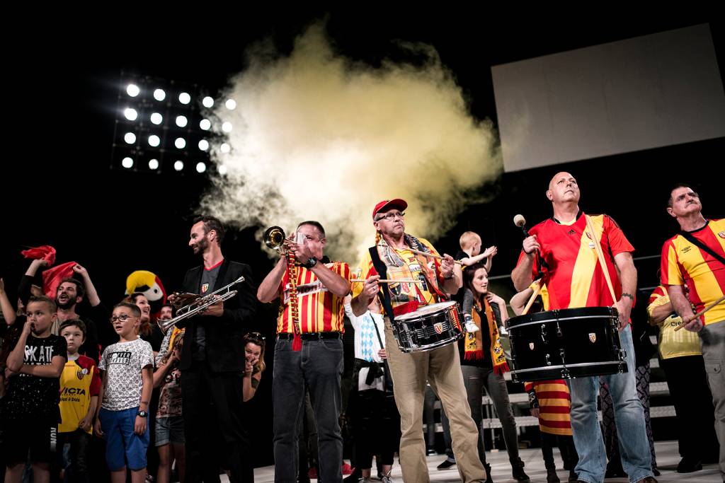Torcedores tocam instrumentos em grupo no palco. Usam camisetas vermelhas e amarelas