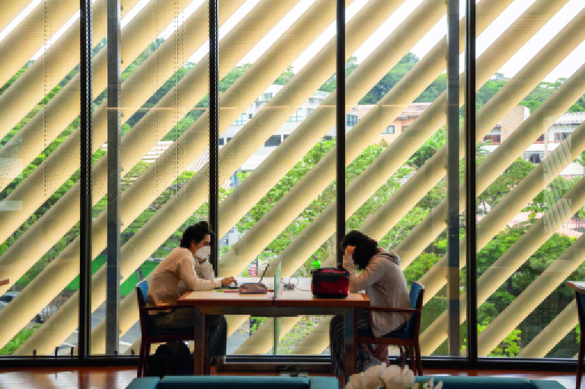 Duas estudantes sentadas em uma mesa de uma das cafeterias do prédio. A parede permite que frestas de luz passem, o que deixa o ambiente aconchegante