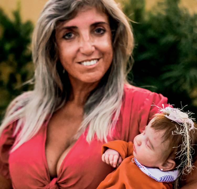 Silvana Rosa, a "cegonha" de Peruíbe, posa com roupa vermelha de decote e boneca realista no colo. Sorri e tem cabelos loiros longos.