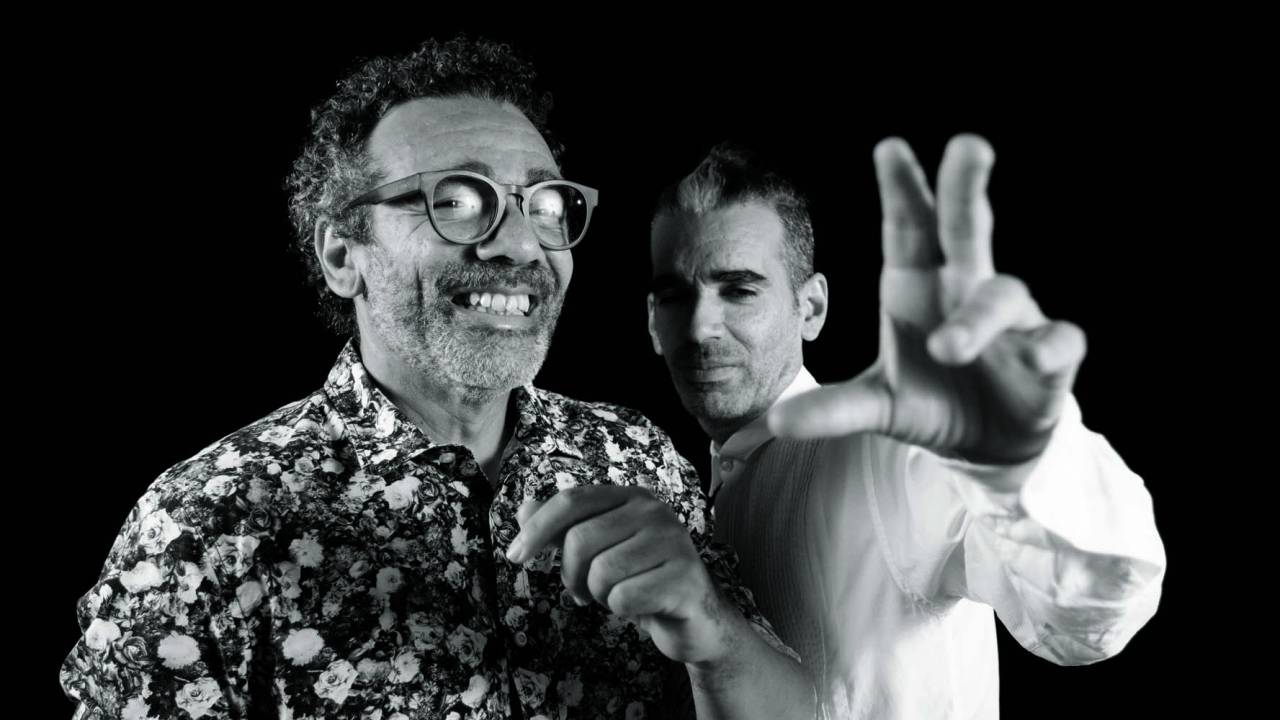 Imagem em preto e branco mostra dois homens. O da esquerda sorri, e o da direita levanta a mão em direção à câmera