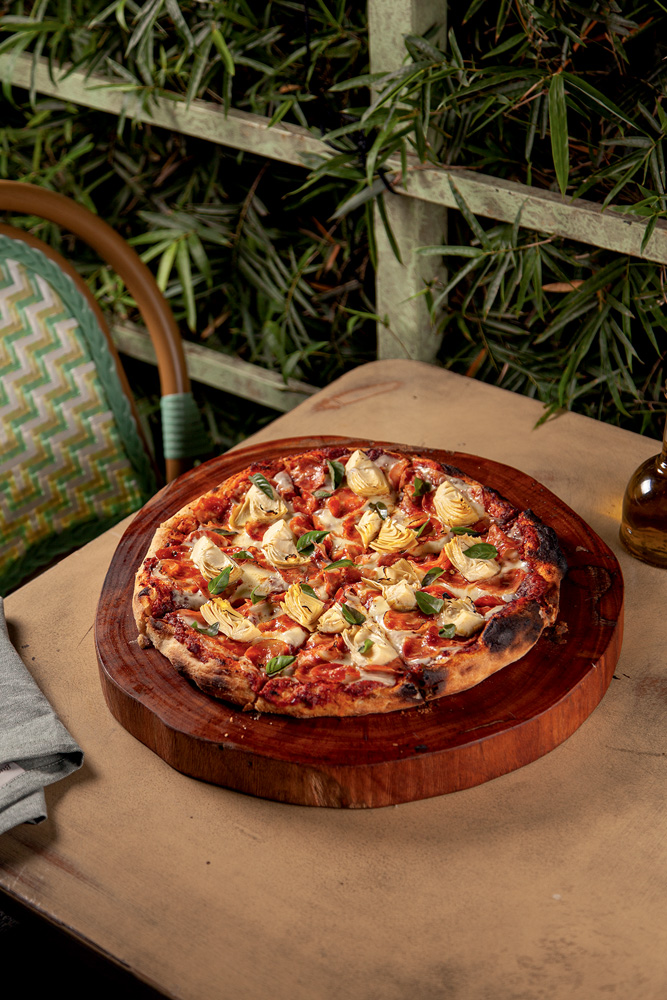 Imagem mostra pizza sobre suporte de madeira, em cima de mesa. Ao fundo, folhas e uma cadeira verde.