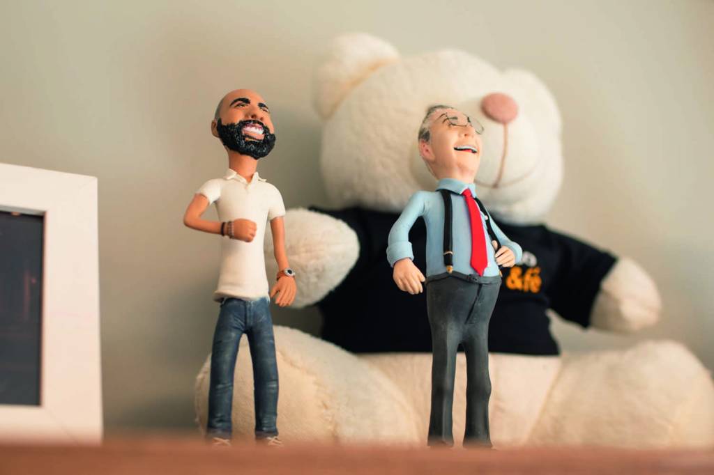 Miniaturas de Bruno e Mário Covas em cima de uma estante, com um ursinho branco de pelúcia atrás