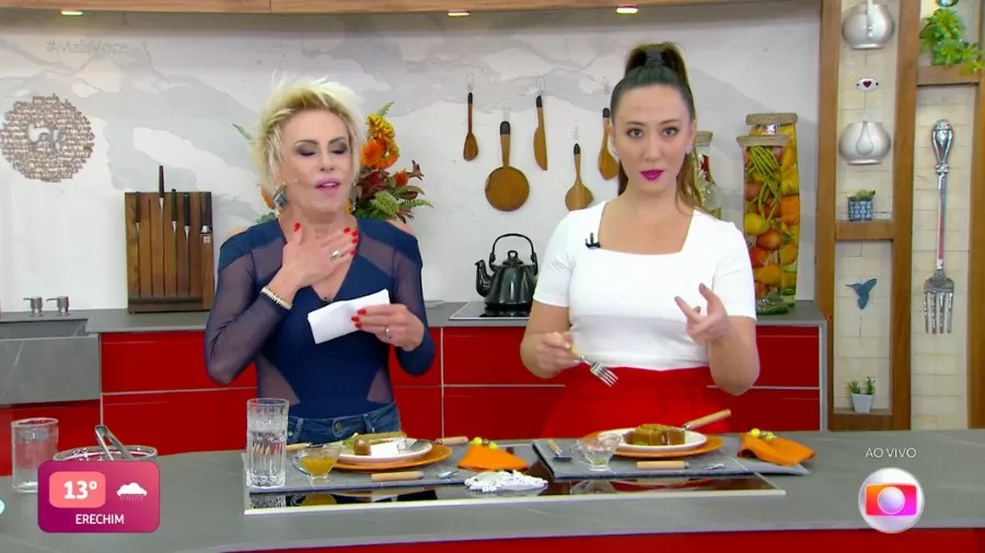 Imagem mostra duas mulheres, uma loira, à esquerda, e uma morena, a direita, atrás de bancada com pratos de comida