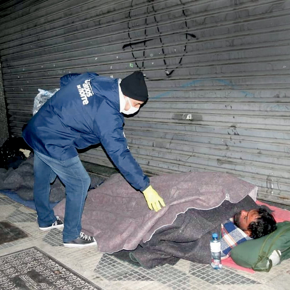 Um homem de roupa de frio e gorro coloca um cobertor cinza em cima de um homem que dorme na rua