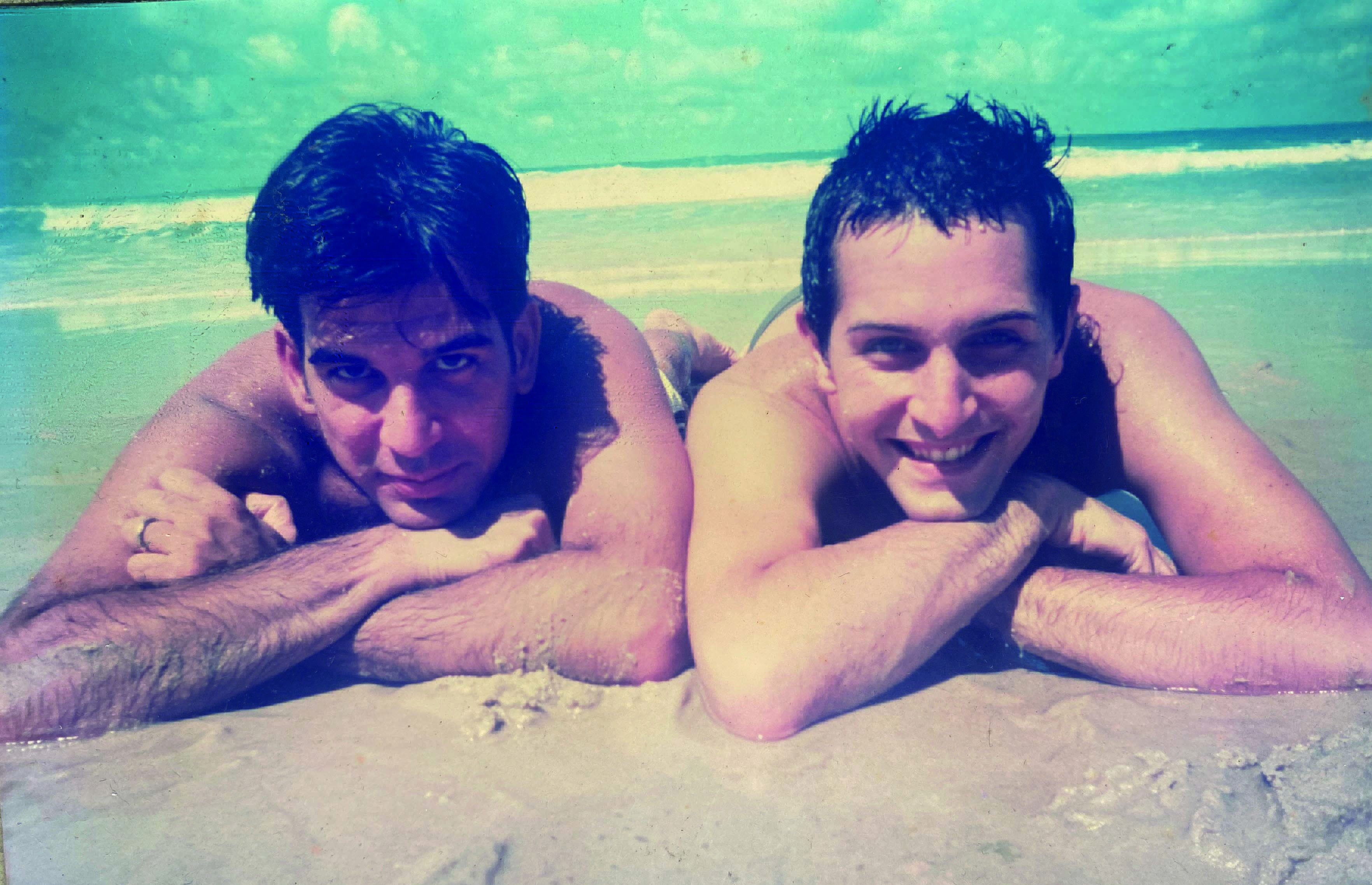 Fábio e Arlindo deitados na areia da praia posam um ao lado do outro para a foto, com a cabeça apoiada nos braços cruzados