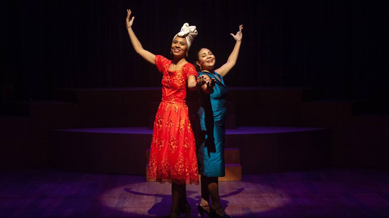 Duas atrizes negras interpretam Carmen Costa e Elizeth Cardoso, cantoras da Era do Rádio. Ambas são negras. Bárbara (Carmen) usa um vestido comprido vermelho e um turbante branco na cabeça. Analu (Elizeth) usa um coque baixo e um vestido longo azul brilhante. Ela estão de costas uma para a outra, sorriem e levantam os braços em sinal de agradecimento