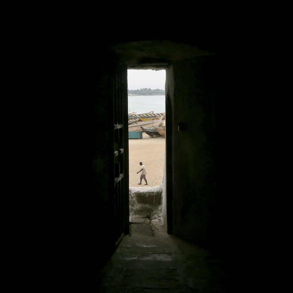 Imagem mostra fresta de porta mostrando praia com um homem caminhando na areia