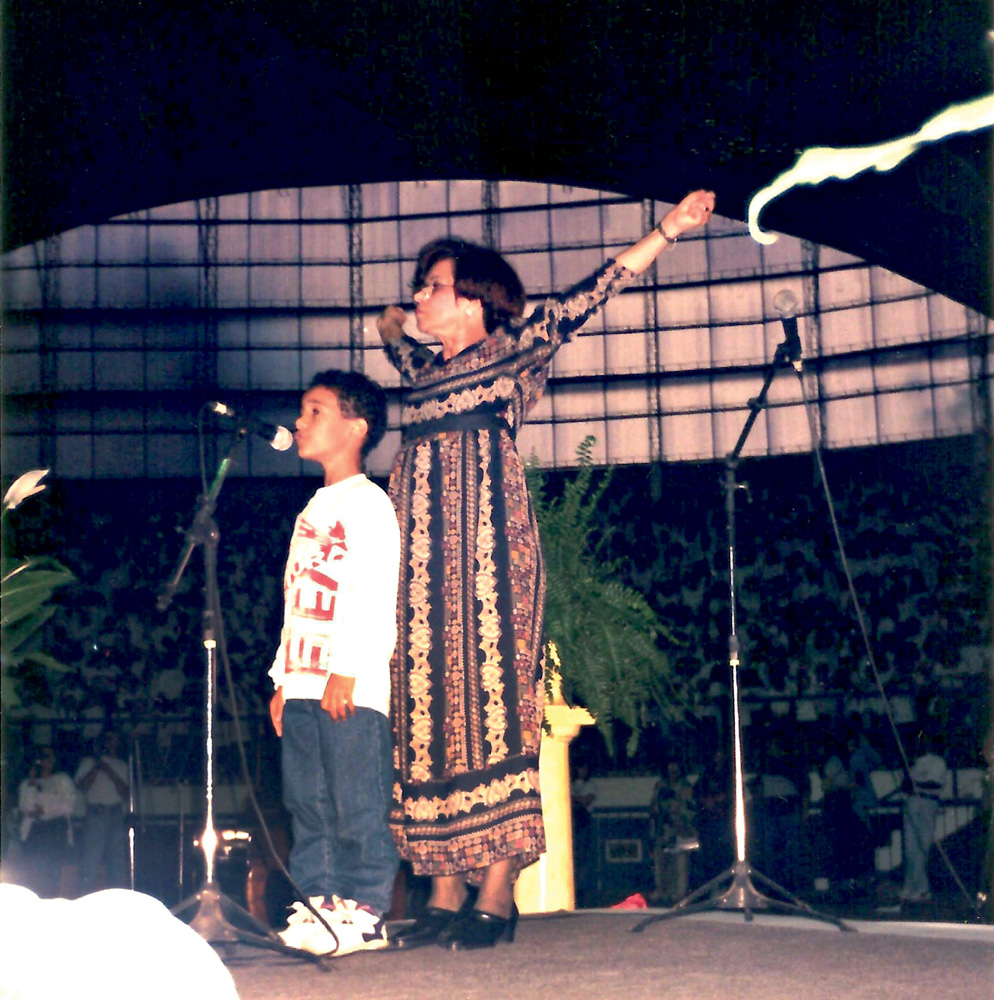 Imagem mostra criança ao lado de mulher com os braços levantados, em cima de palco, cantando em microfone