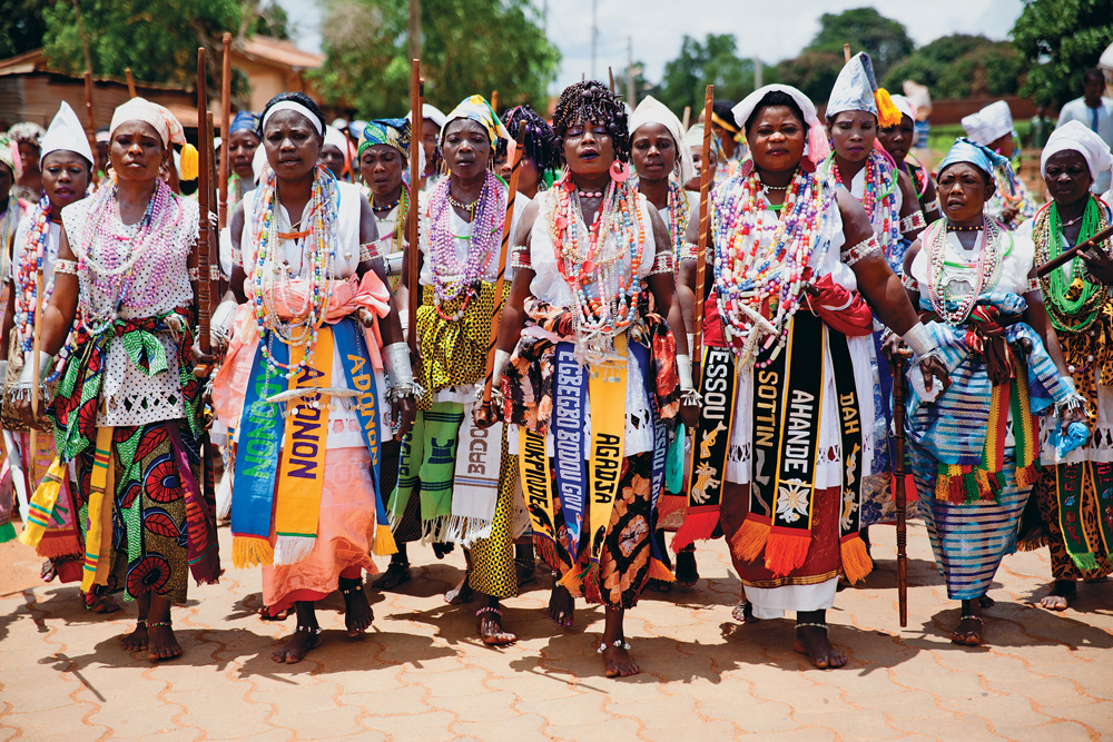 Imagem mostra mulheres caminhando em chão de terra, vestindo saias e roupas com diversos adereços coloridos