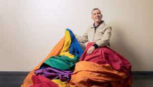 ABRE lgbtRENATOVITERBO5 – crédito Leo Martins.jpg Imagem mostra homem segurando bandeira LGBT grande, sentado, sobre suas pernas.
