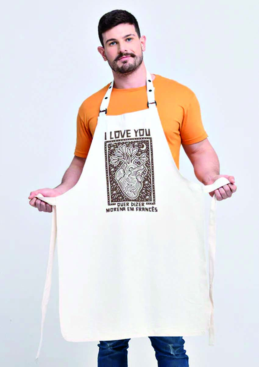 Modelo branco e magro usa avental bege com desenho de cora&ccedil;&atilde;o em estilo de cordel e a frase 