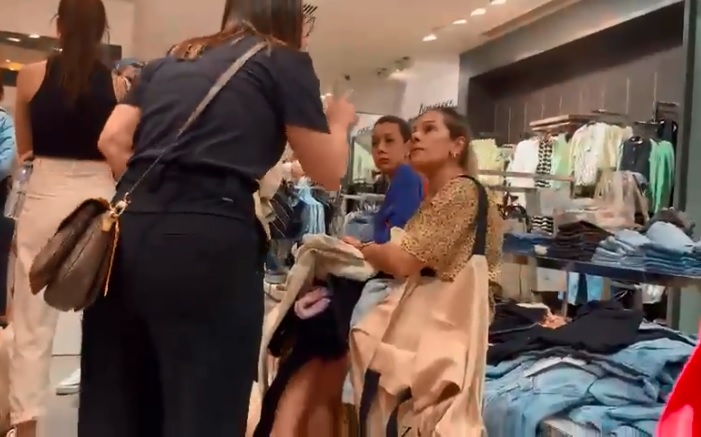 Frame de vídeo com duas mulheres brigando entre araras de roupa.