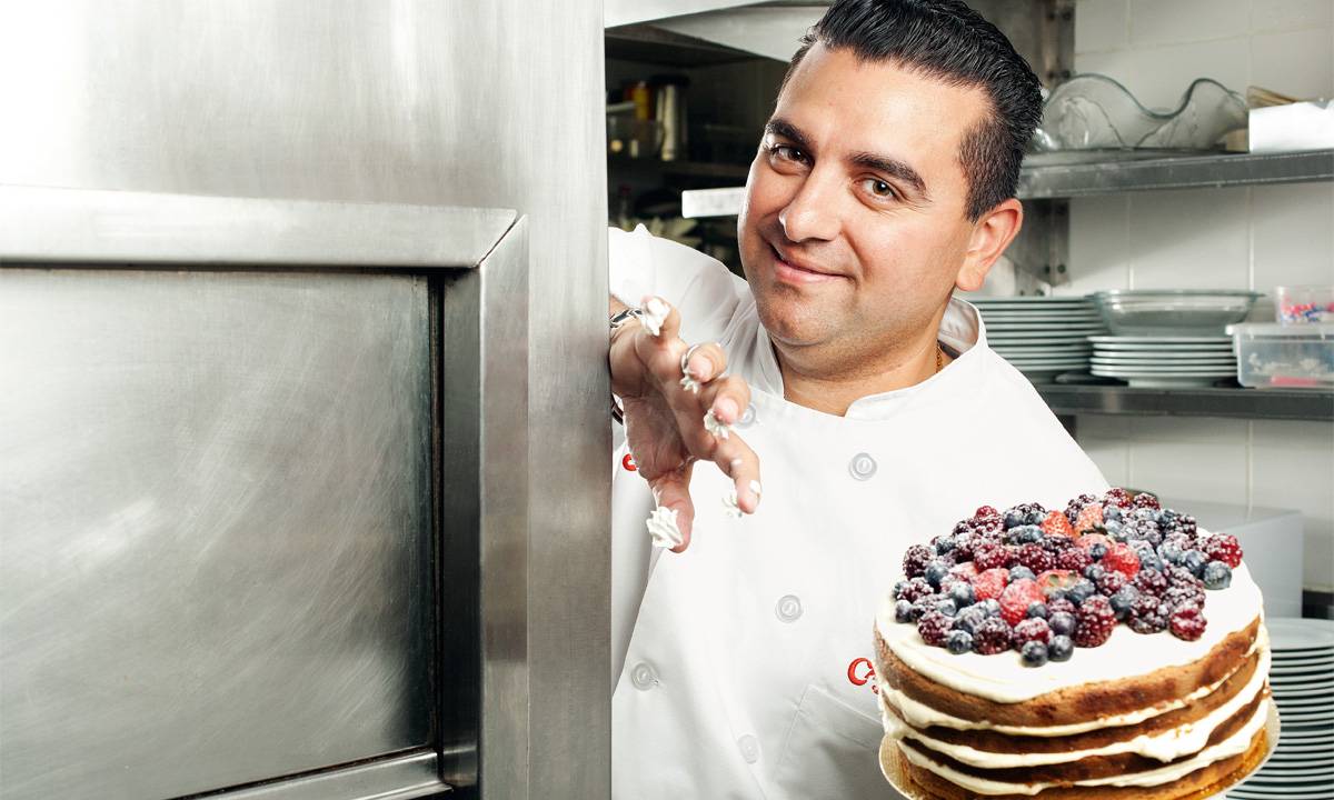 Buddy Valastro posa em ambiente de cozinha enquanto segura um bolo coberto por frutas vermelhas