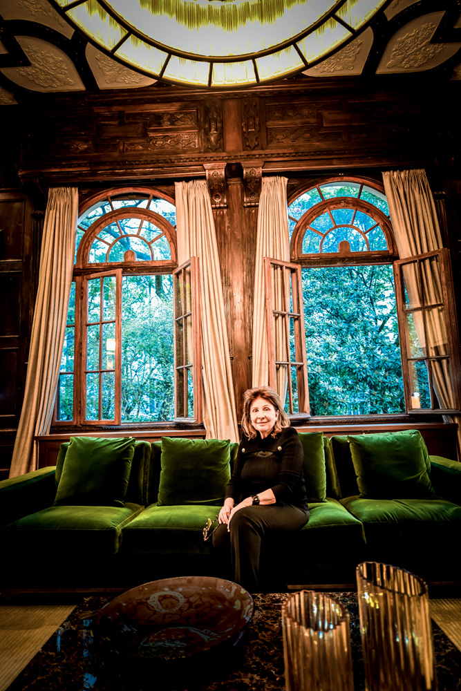 Luciana, arquiteta de interiores, sentada em um luxuoso sofá verde oliva brilhante no Casarão. Atrás há duas grandes janelas com cortinas que voam com o vento. Ela é uma senhora idosa, branca e magra, de cabelos curtos e loiros. Usa uma roupa inteiramente preta.