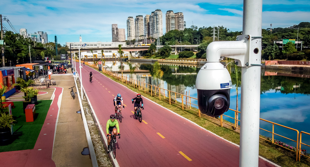 Em foco, câmera de inteligência artificial instalada na ciclovia do Rio Pinheiros e ciclistas pedalando ao fundo