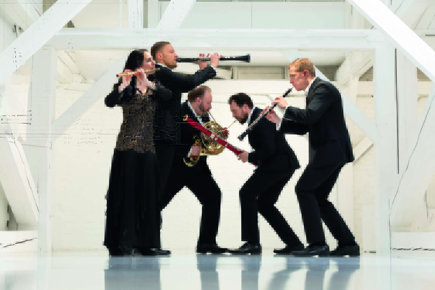 Imagem mostra cinco pessoas com roupas de gala tocando instrumentos de sopro, em pé.