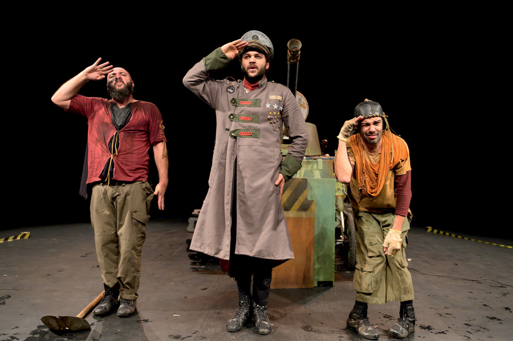 Três atores homens usam roupa de soldado e prestam continência um ao lado do outro. Ao fundo, há um tanque de guerra cenográfico