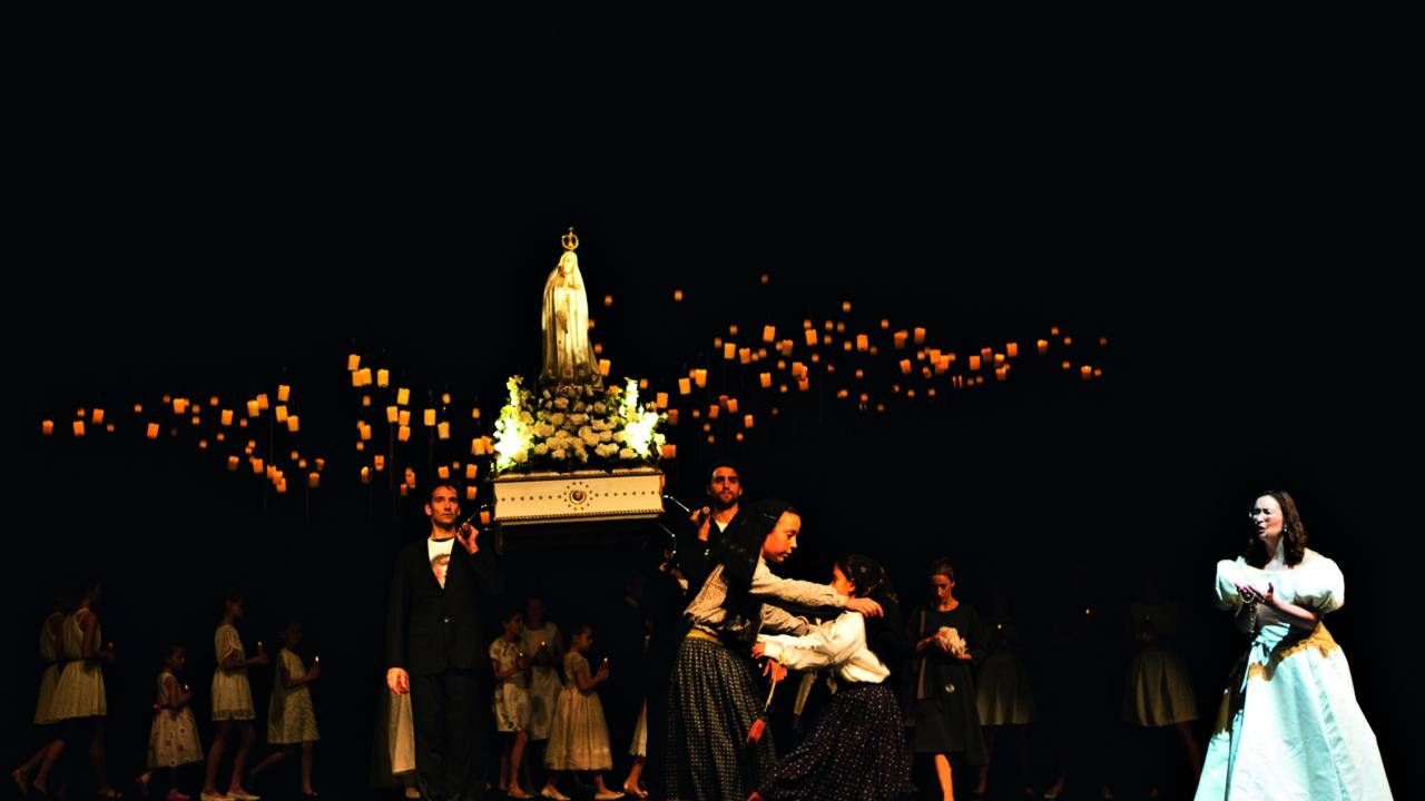 Dançarinos no palco cheio de luzes amarelas. Dois seguram uma imagem religiosa da santa