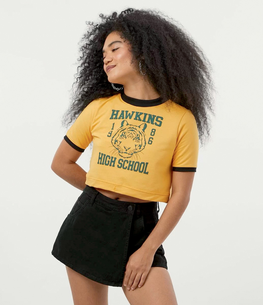 Modelo negra, alta e magra, de cabelos crespos compridos, usa cropped amarelo com a estampa de Hawkins High