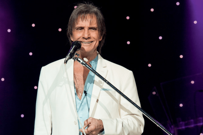 Imagem mostra homem de terno branco e camisa azul clara, sorrindo, em frente a um microfone
