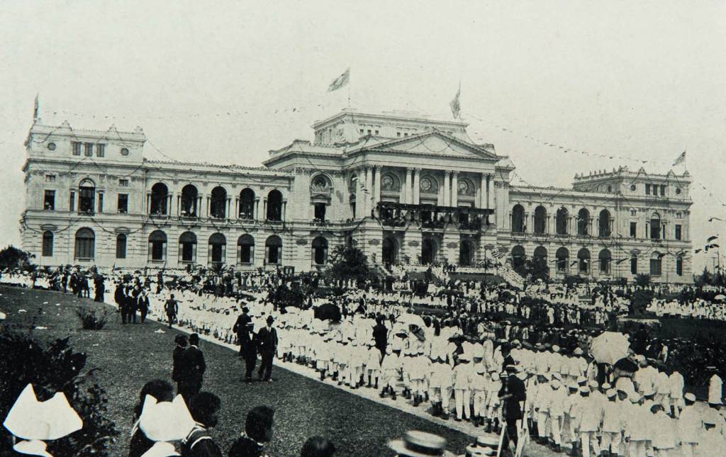 Imagem em preto e branco mostra multidão ao redor de jardim de edifício clássico.