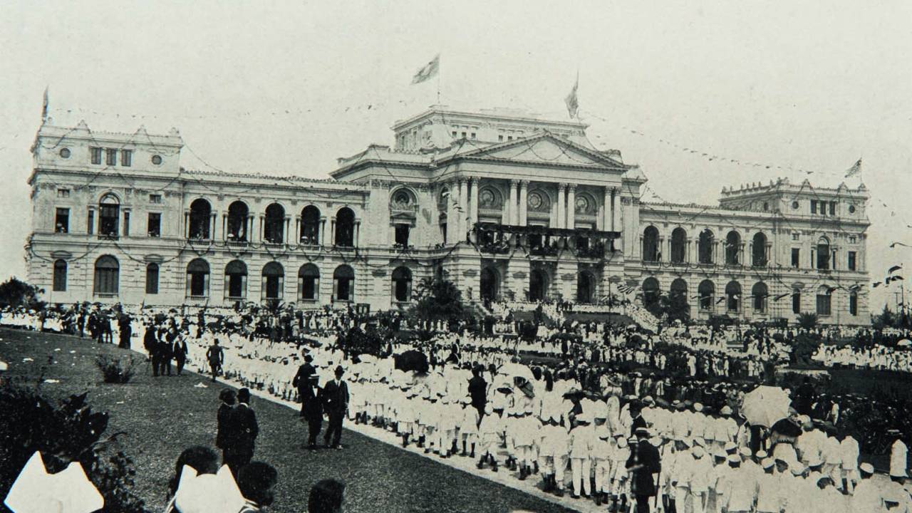 Imagem em preto e branco mostra multidão ao redor de jardim de edifício clássico.