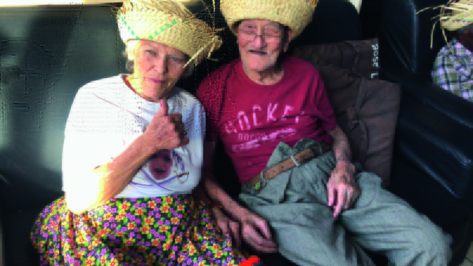 Um senhor japonês e uma senhora de cabelos brancos e olhos azuis vestem chapéis de festa junina sentados em um sofá