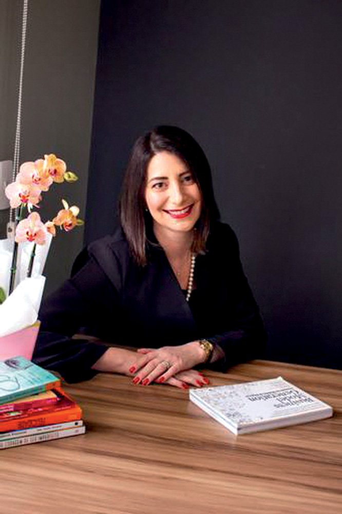 Isabela Nassif aparece sorrindo sentada a uma mesa e ao seu lado esquerdo há um buquê de flores rosas e laranjas