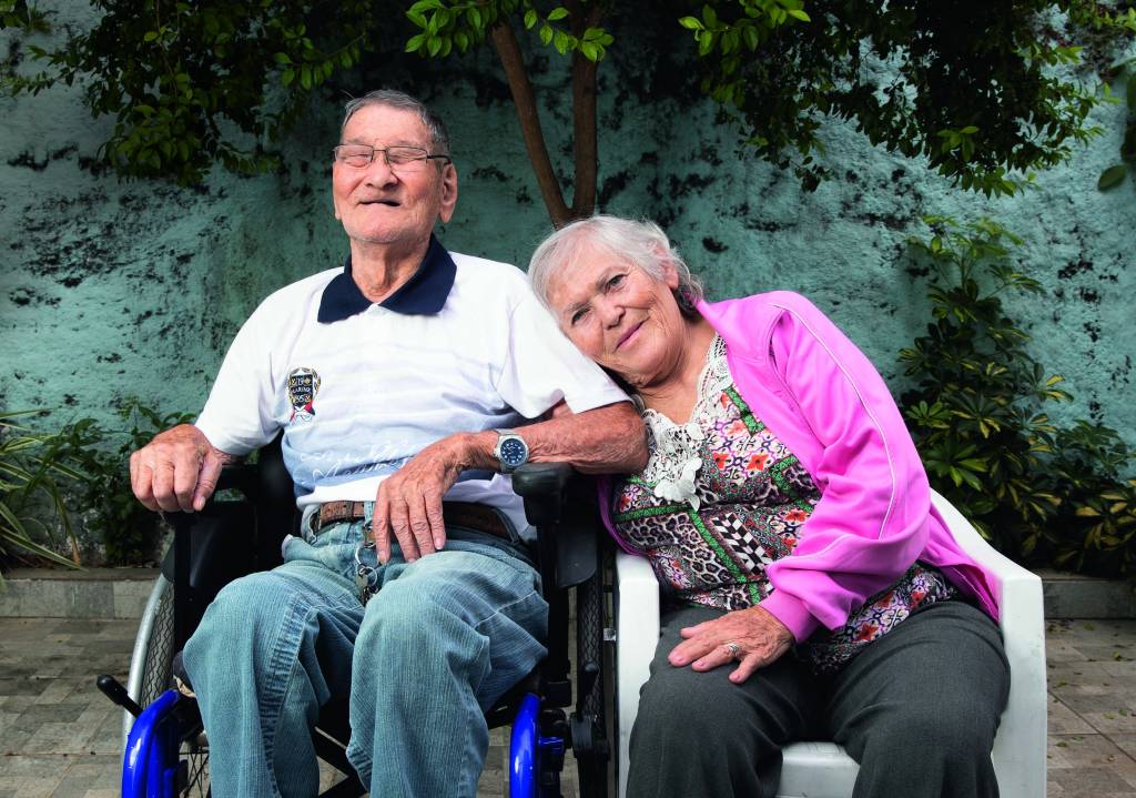 Um senhor japonês está sentado em uma cadeira de rodas ao lado de sua namorada, uma senhora de cabelos brancos e olhos azuis sentada em uma cadeira branca. Eles estão em um jardim e ela encosta a cabeça no ombro dele