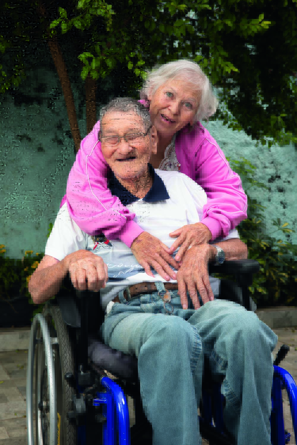 Um senhor japonês está sentado em uma cadeira de rodas ao lado de sua namorada, uma senhora de cabelos brancos e olhos azuis que se apoia na cadeira e encosta o rosto próximo ao seu. Eles estão em um jardim