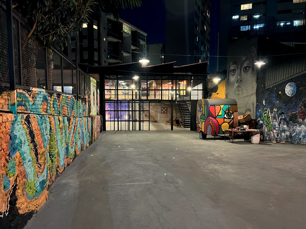 Foto exibe galpão de chão cinza, food truck no fundo e janelões de vidro, com grafite colorido na parede à esquerda.