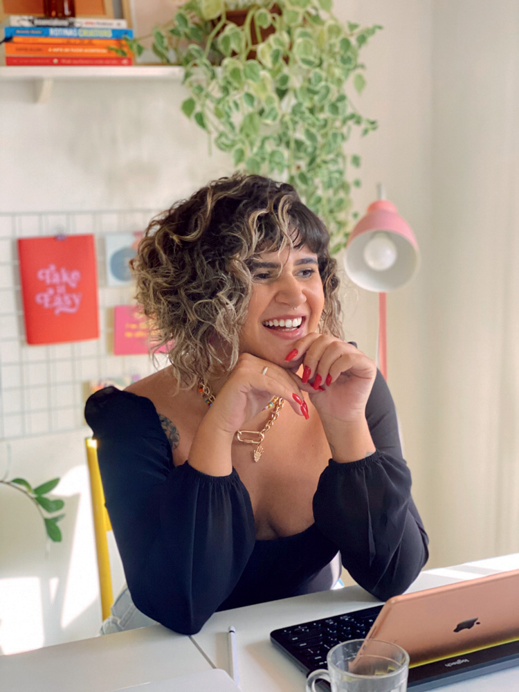 Larissa aparece com os dois braços apoiados em uma mesa e sorrindo, com a mão esquerda encostado no rosto