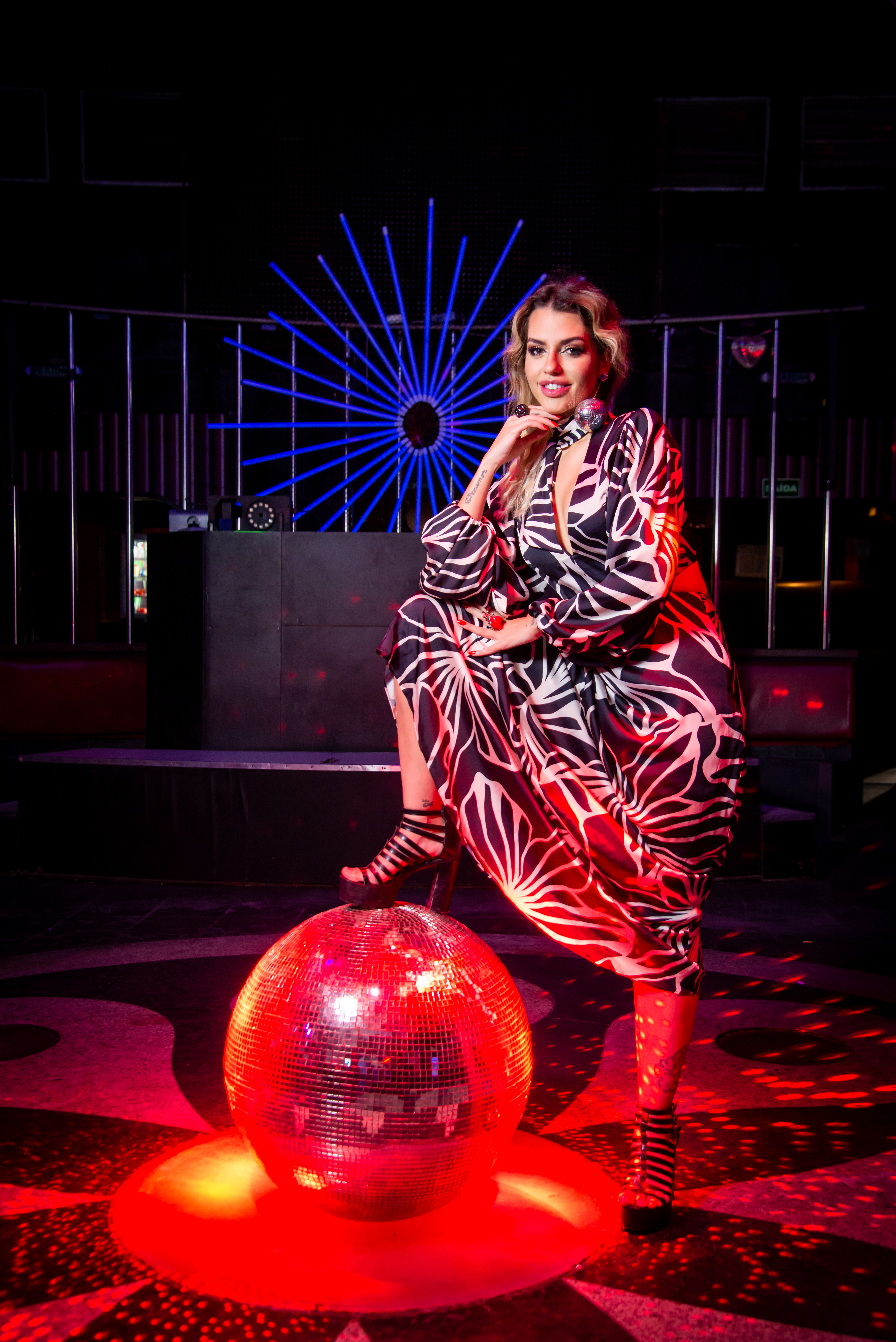 Lili Scott posa com uma perna apoiada em bola de discoteca iluminada com luz vermelha. Ela veste um longo vestido estampado e está com uma das mãos no queixo.