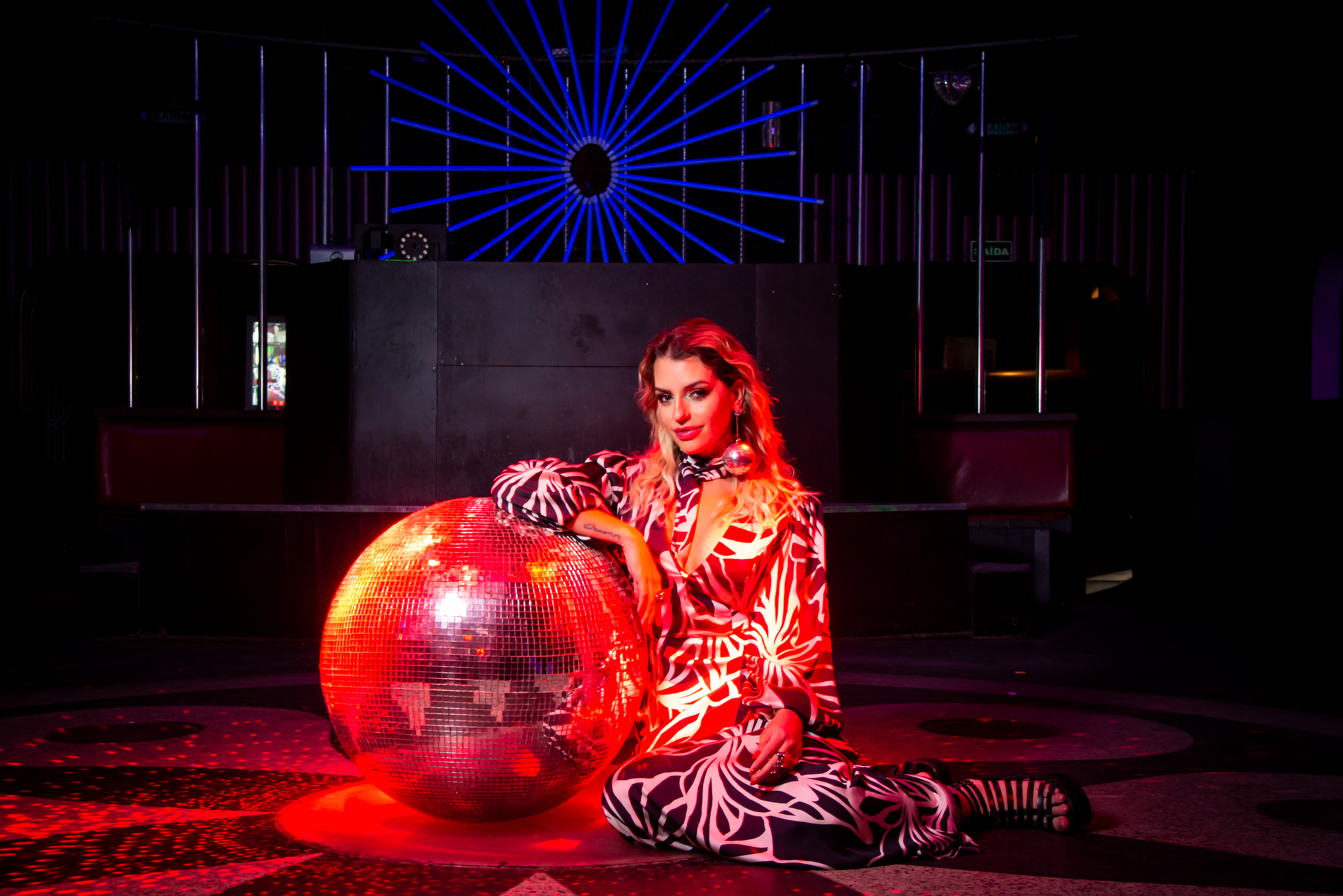 Lili Scott posa sentada e apoiada em bola de discoteca iluminada com luz vermelha. Ela veste um longo vestido estampado e está com um dos ombros encostado na bola.