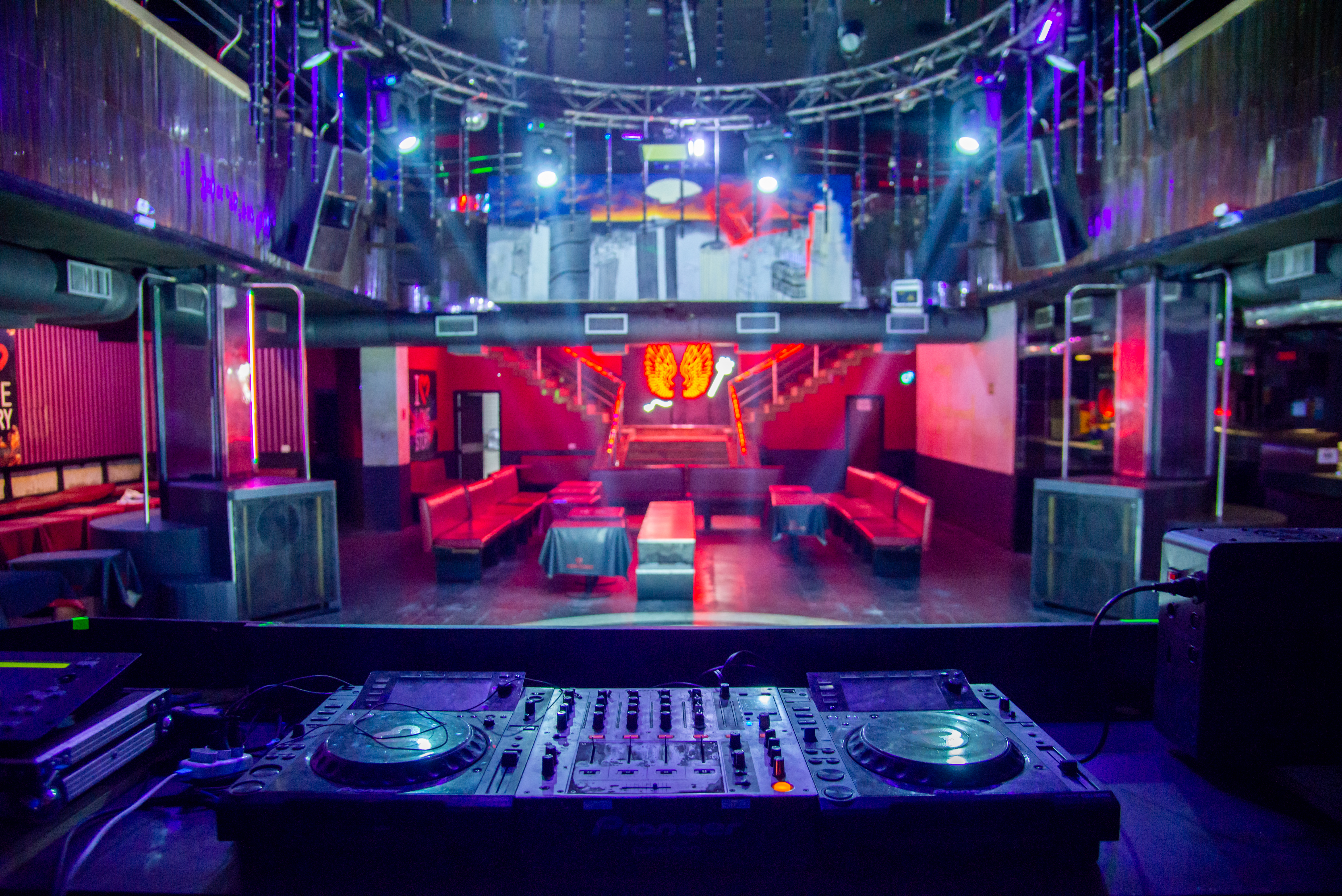 Foto exibe cabine do DJ e pista com sofás vermelhos e iluminação à frente.