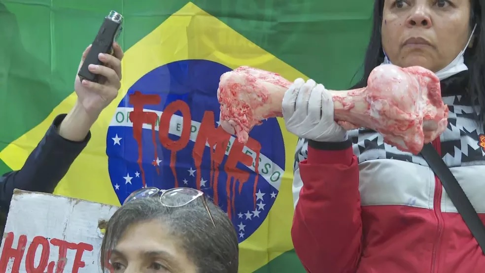 Mulher segura osso avermelhado com luva em frente a bandeira do Brasil com a palavra FOME escrita em tinta vermelha, simulando sangue.