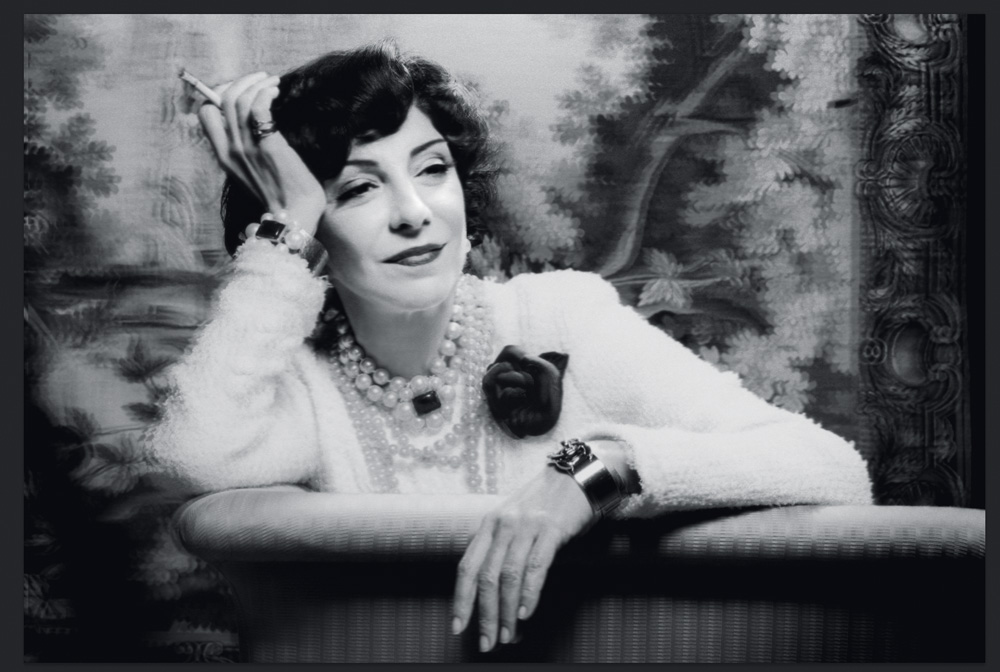 Imagem em preto e branco mostra mulher sentada em sofá, com um cigarro na mão.