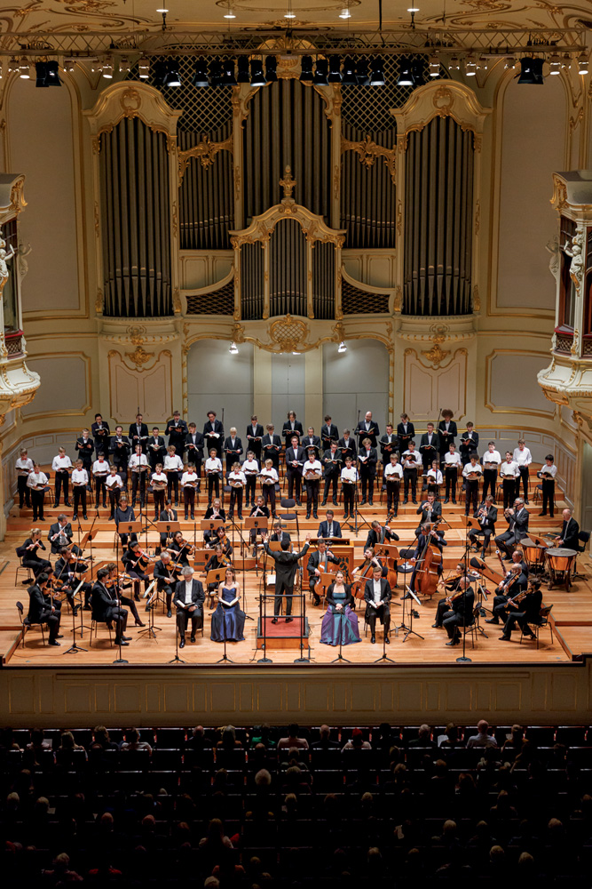 Imagem mostra sala de concertos com orquestra e coro de crianças no palco