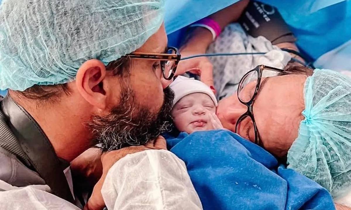 Foto de Juliano Cazarré ao lado da filha recém-nascida e a esposa. Vestem roupas cirúrgicas.