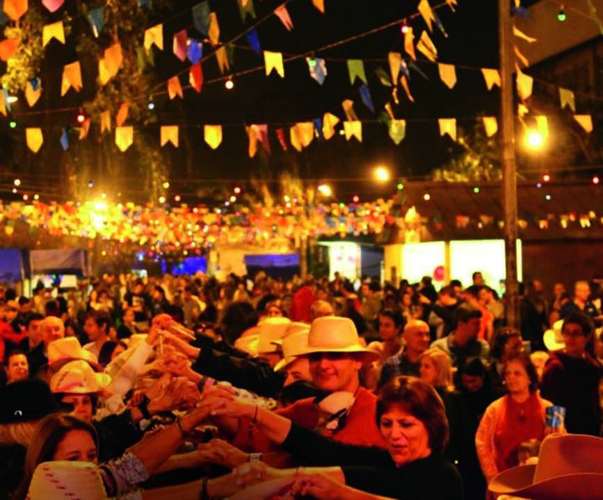 Imagem mostra diversas pessoas com roupas de festa junina, a noite, dançando quadrilha
