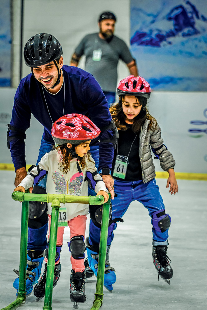 Menina pequena patina no gelo segurando em um andador verde. O pai se apoia junto com ela. Atrás, outra menina patina sem andador