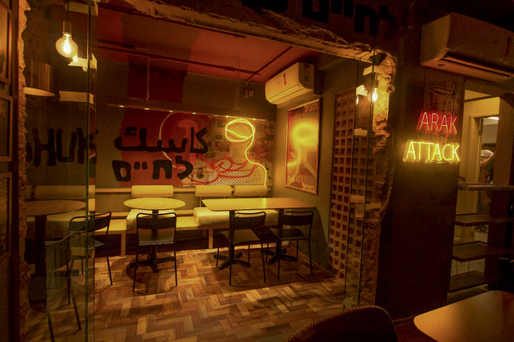 Salão do Shuk, restaurante árabe, com mesas e cadeiras brancas. Um vidro separada o ambiente da frente e o ambiente das cadeiras, que fica atrás. No vidro está escrito palavras em hebraico e árabe. O ambiente é escurinho, iluminado por LEDs amarelas e vermelhas