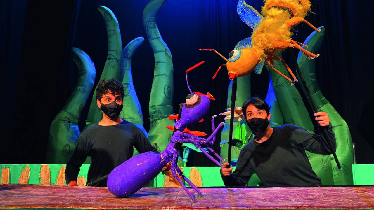 Dois atores vestidos de preto manipulam um boneco de abelha e outro de formiga no palco