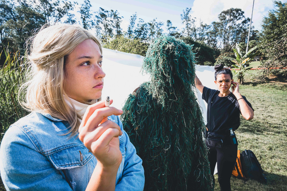 Atriz com peruca loira fuma um cigarro em um set de filmagens ao ar livre. Ao fundo, uma mulher interpreta uma funcionária da produção do filme e um ator usa um figurino de monstro de pântano