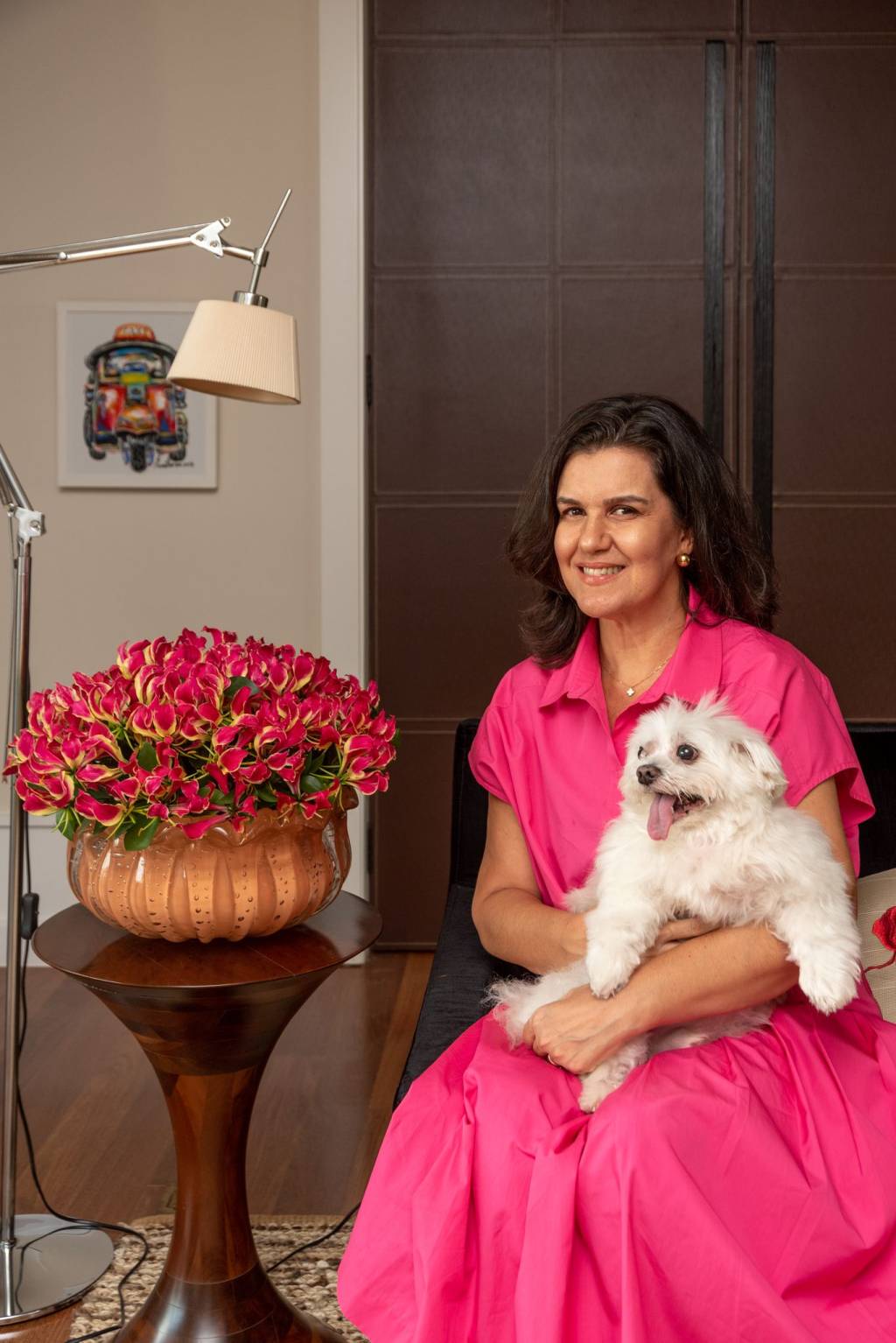 Tetê Castanha posa sentada com cachorrinho de pelos brancos ao lado de arranjo rosa. Veste vestido rosado também.