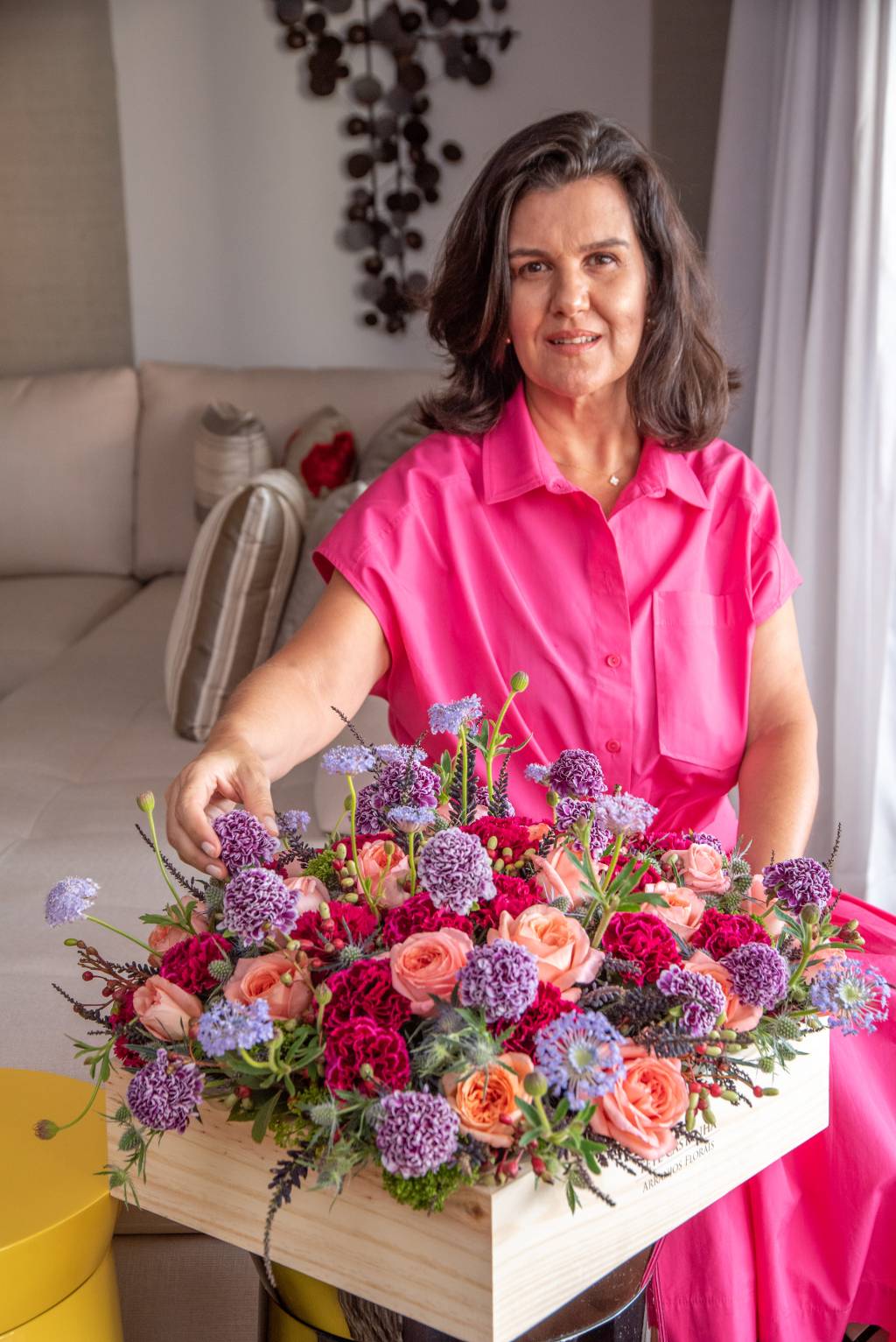 Tetê Castanha posa vestida de rosa mexendo em arranjo floral com as cores vermelho, roxo e rosa.