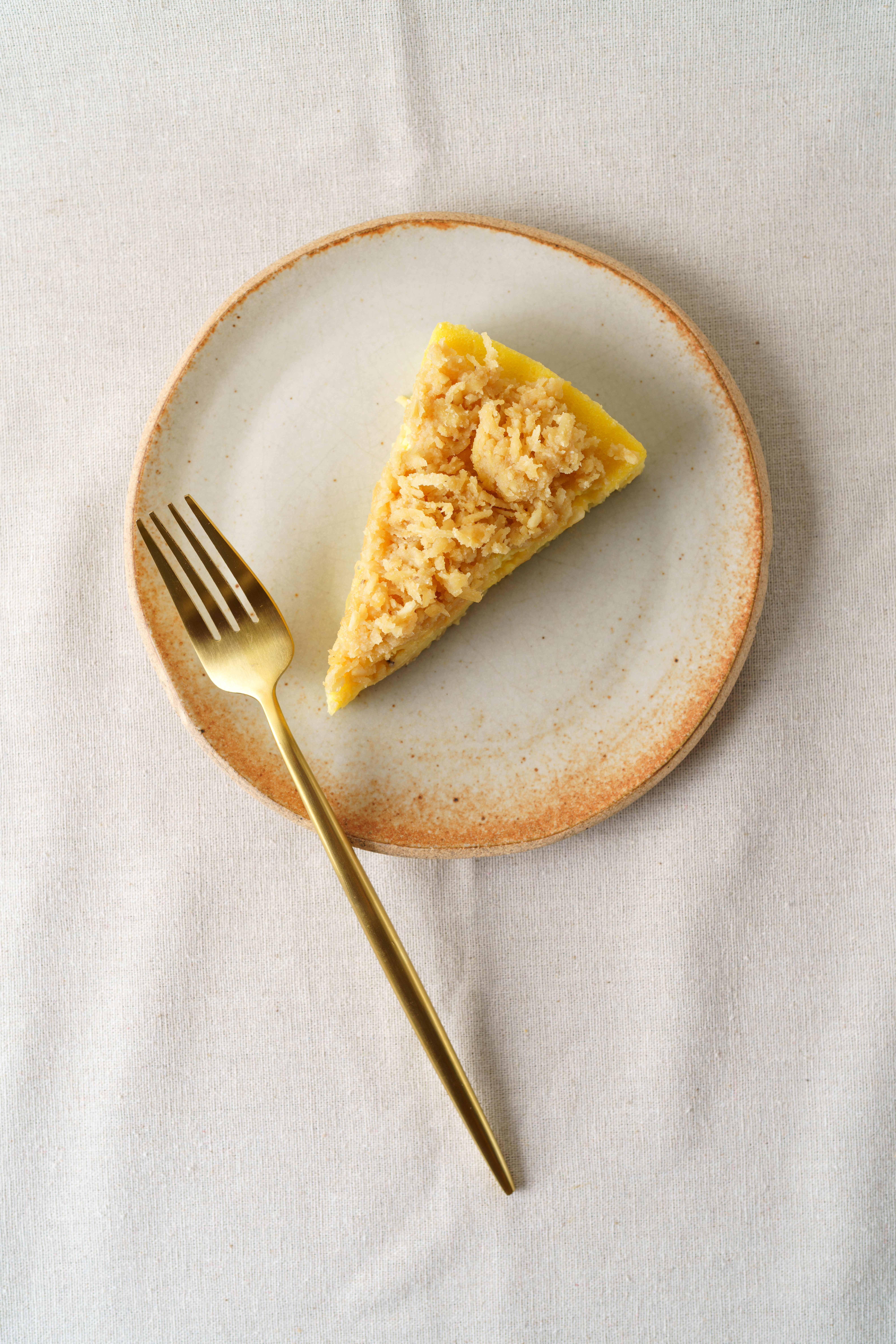 Torta de cor amarelada coberta por pedaços de coco queimado servida em prato de cerâmica junto de um longo garfo